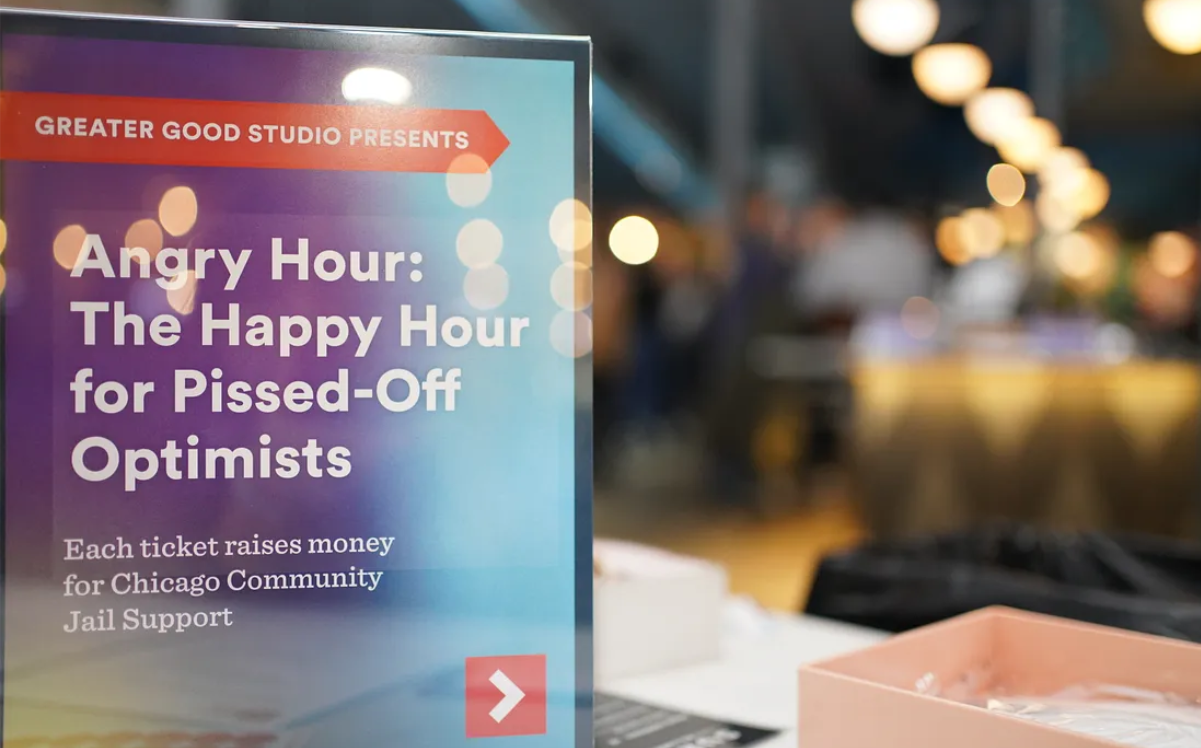 A sign on the left reads “Greater Good Studio Presents Angry Hour: The Happy Hour for Pissed-Off Optimists. Each ticket raises money for Chicago Community Jail Support.” Blurred lights and shapes in the background on the right.”