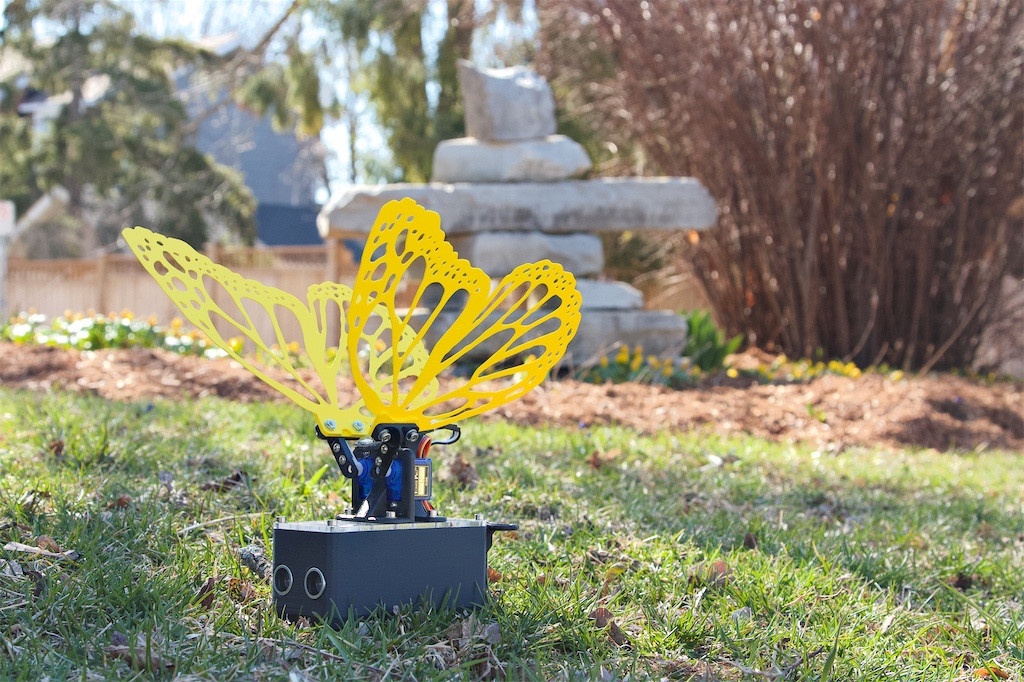 Robot Butterfly with yellow wings in the foreground. It's a beautiful fresh and bright spring day, with light streaks and shadows cast over the short and slightly green grass. In the background is an Inunnguaq sculpture, and at the feet are yellow sprouts