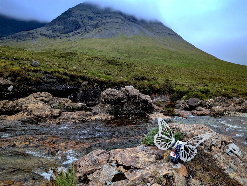 Robot Butterfly with white translucent wings perched on a rock by a stream, in front of a large mountain, partially shrouded in clouds