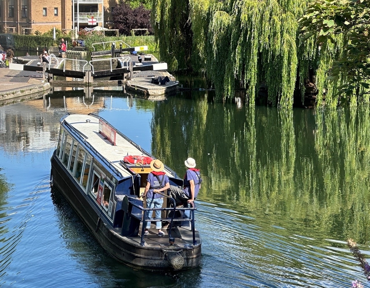 A dark blue canal boat moves through calm green water toward a wooden lock gate. Two people wearing sun hats and life jackets stand at the stern. A large, lush weeping willow hangs over the water on the right