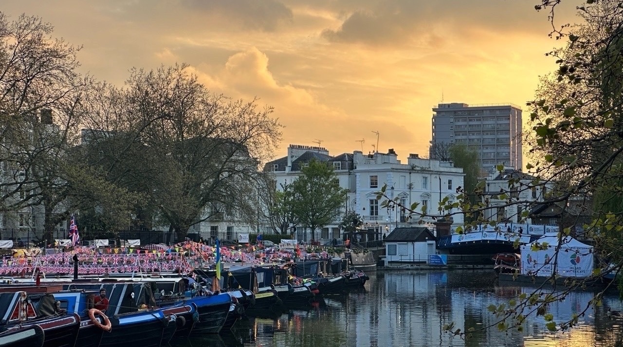 A wide view of the canal at Little Venice during golden hour. Numerous narrowboats are moored closely together, heavily decorated with Union Jack bunting and colourful flags. In the background, white Regency-style buildings and a modern high-rise.
