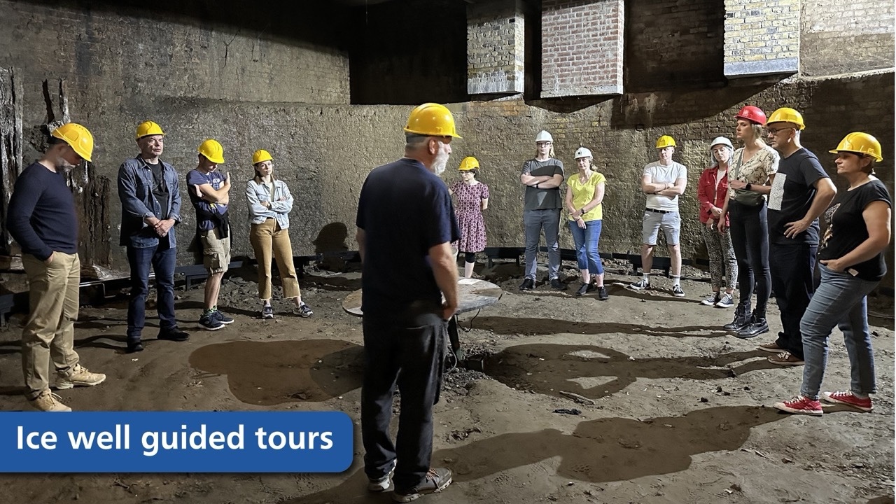 A group of twelve visitors in casual clothes and yellow safety helmets stand in a semi-circle inside a dimly lit, vast subterranean chamber. They are listening to a guide, seen from behind, who stands in the centre of the dirt floor. The curved walls are