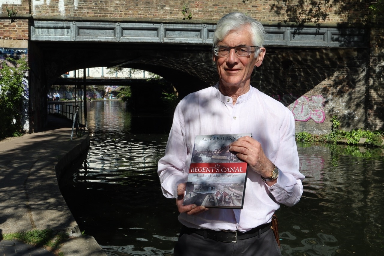 Charlie, a man with grey hair and glasses, wearing a light pink shirt. He stands by a towpath, holding a copy of the book 'The Regent’s Canal'. In the background, a dark brick bridge arches over the still water of the canal