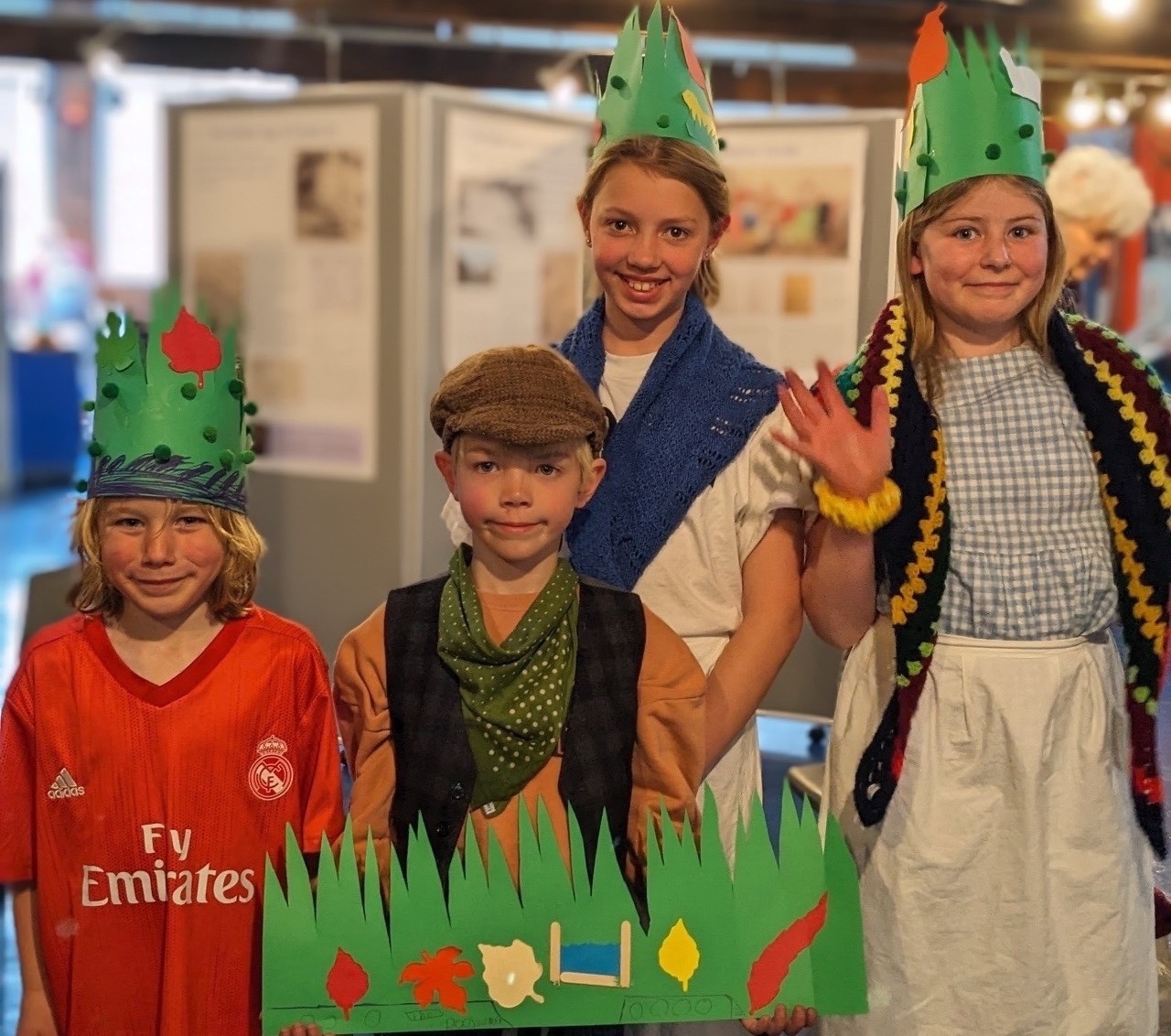 Four smiling children stand in a museum gallery. Three wear green paper crowns decorated with leaves, while one boy wears a flat cap and waistcoat. They hold a large green card cutout featuring autumn leaves and a drawing of a canal lock.