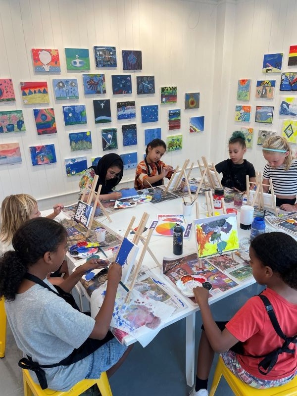 View inside the gallery during an art class. A diverse group of seven children are painting on canvases set up on easels in front of them. All of them look focused and engrossed.