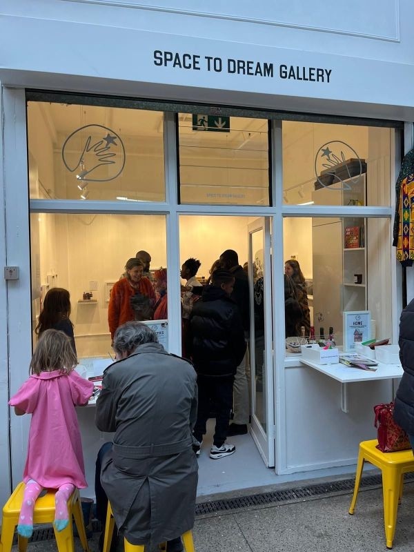 View of the gallery from the outside. A man and a young girl are absorbed in a creative activity on one of the tables outside the gallery while visitors inside seem to be looking around and chatting. People are smiling; the gallery looks busy and joyous.