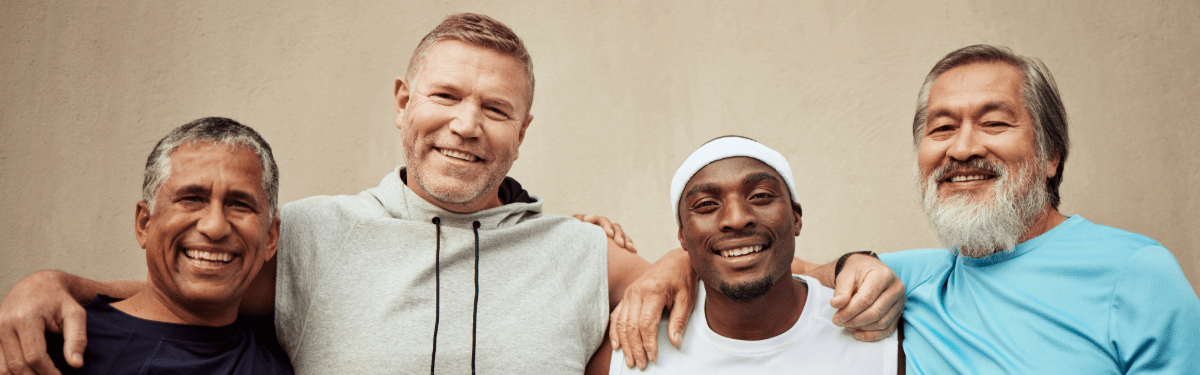 Four men standing close together smiling, arms around each other, wearing casual athletic clothing against a beige wall.