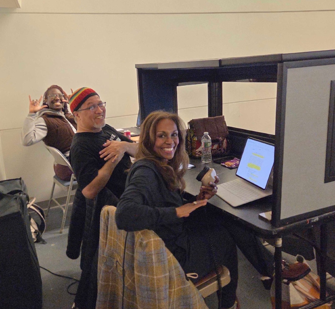Photo of three people sitting at a booth, an African-American woman wearing a gray sweatshirt signing I Love You in ASL, a Latinx male wearing a black shirt and crochet beanie and a South Asian woman wearing a navy top and holding a stenographer mic.