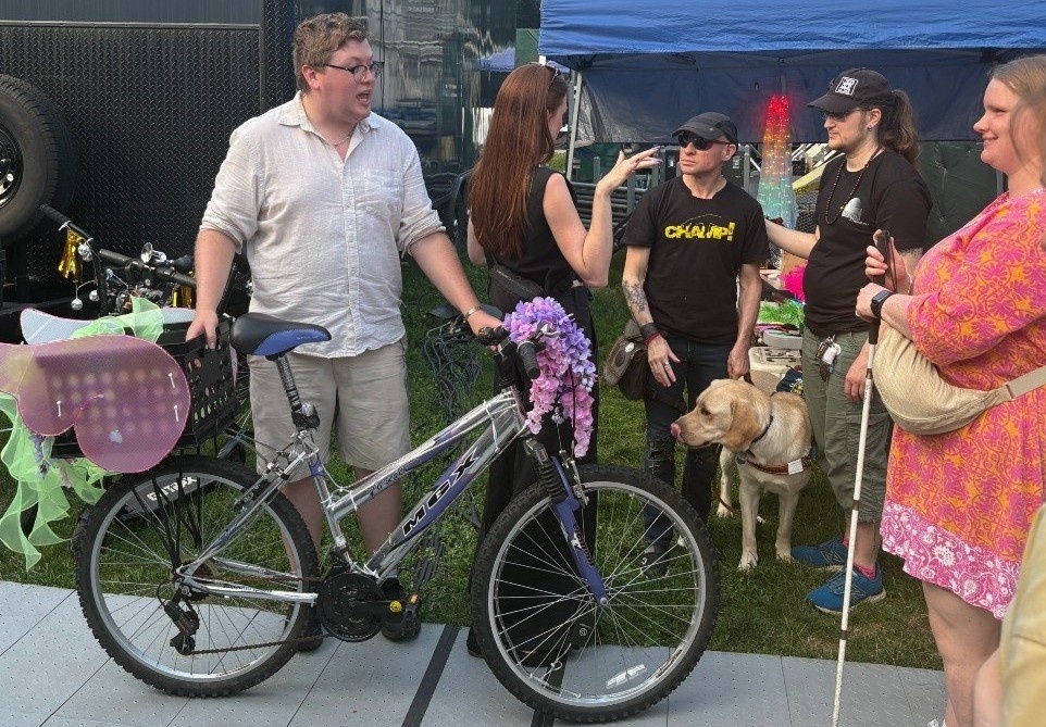 DeafBlind patron Cai and a Blind patron from England receiving a tactile tour and learning about a decorated bike by a CSC production associate and Ingrid interprets in ASL.