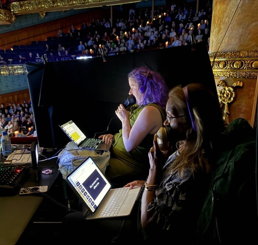 Describers Lali and Gwen sitting in the box seats behind a black curtain; Gwen reading the libretto and in-show AD while Lali reads the English surtitles of the French opera, both speaking into stenographer mics.