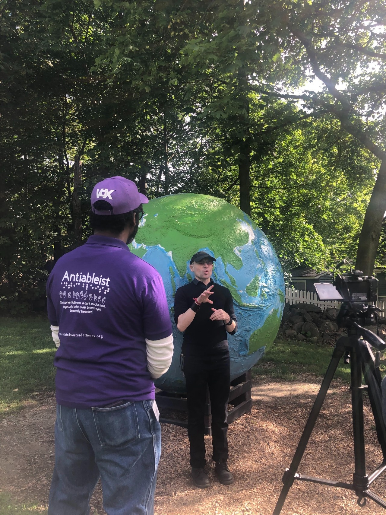DeafBlind expert Cai Steele in front of a statue globe filming for the modules with Christopher Robinson standing near him out of the filming frame wearing a purple Vox shirt.