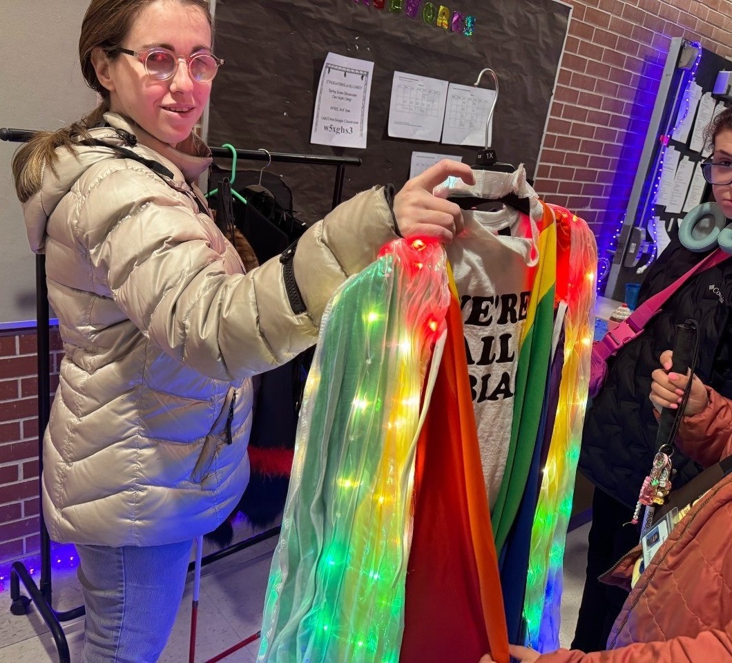 Tanja giving a tactile tour to Perkins students backstage - she is holding the character Angie's rainbow cape with string lights and a white shirt that reads "We're all lesbians."
