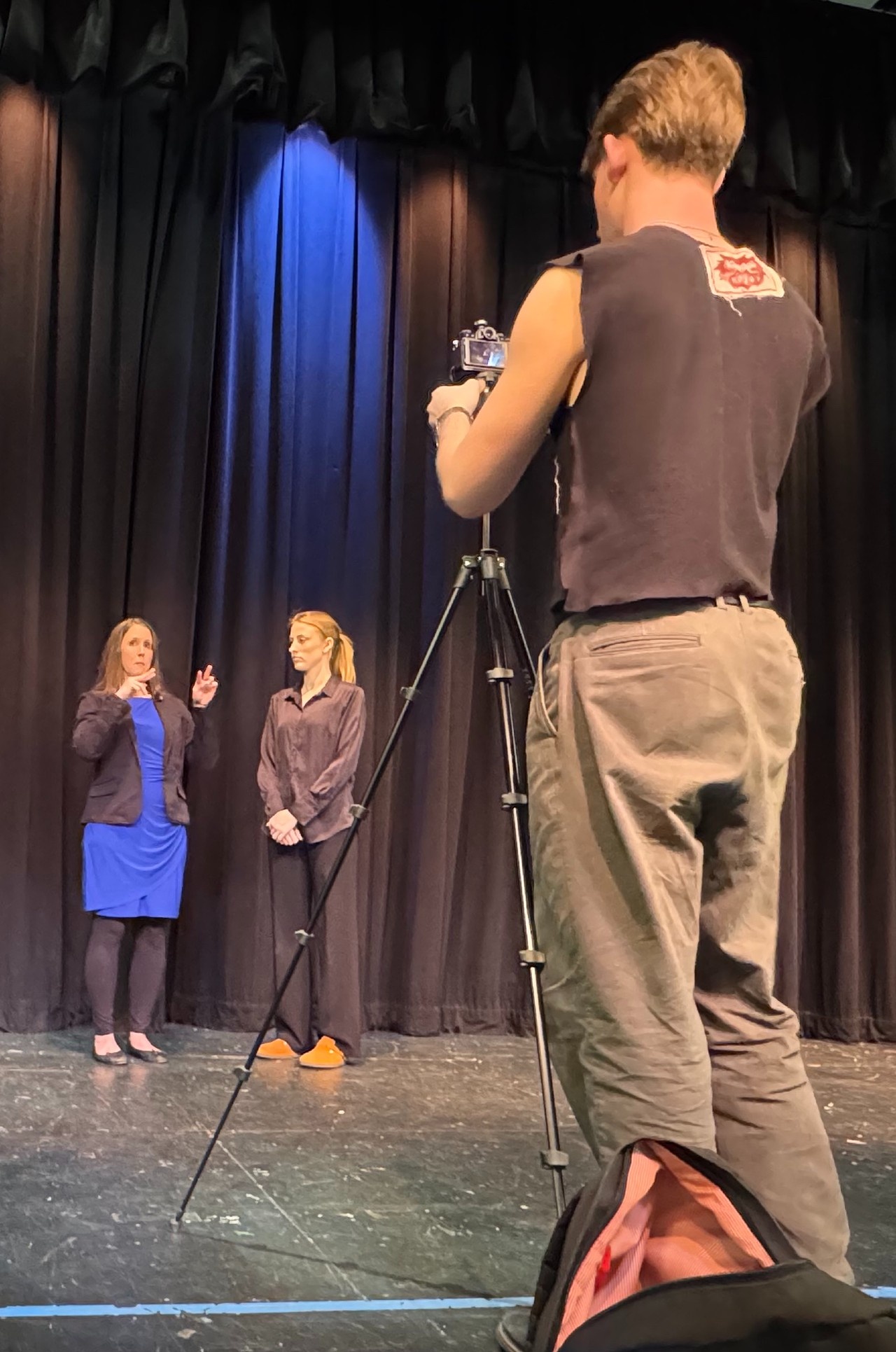 Voxer Connor filming DeafBlind expert Kerry Thompson who is explaining best practices in ASL and English on stage in front of a curtain. Next to her is interpreter Ingrid O'Dell.