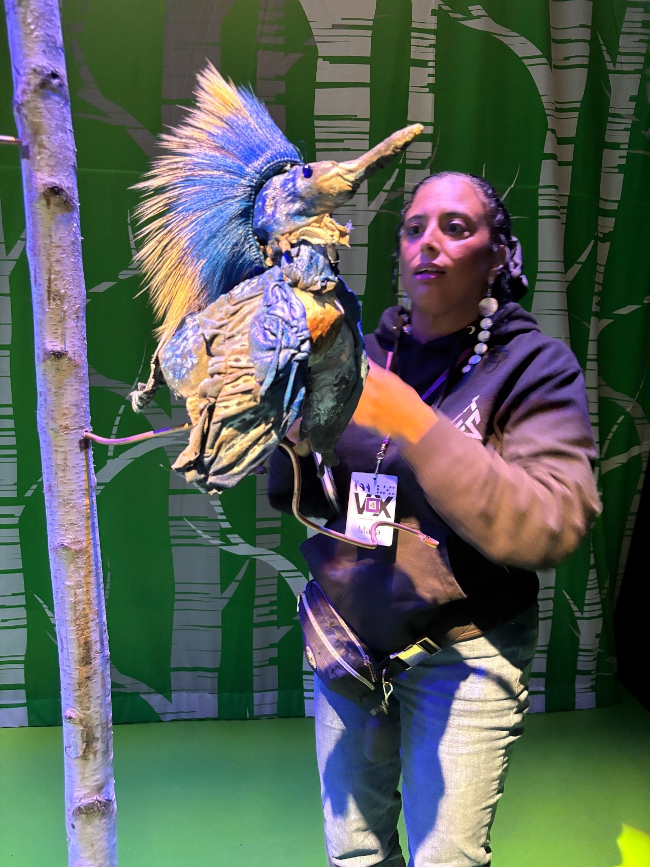 Maria up close and personal with a Kingfisher bird puppet which wears a crest of blue, black and grey porcupine hair, perching on a birch tree.