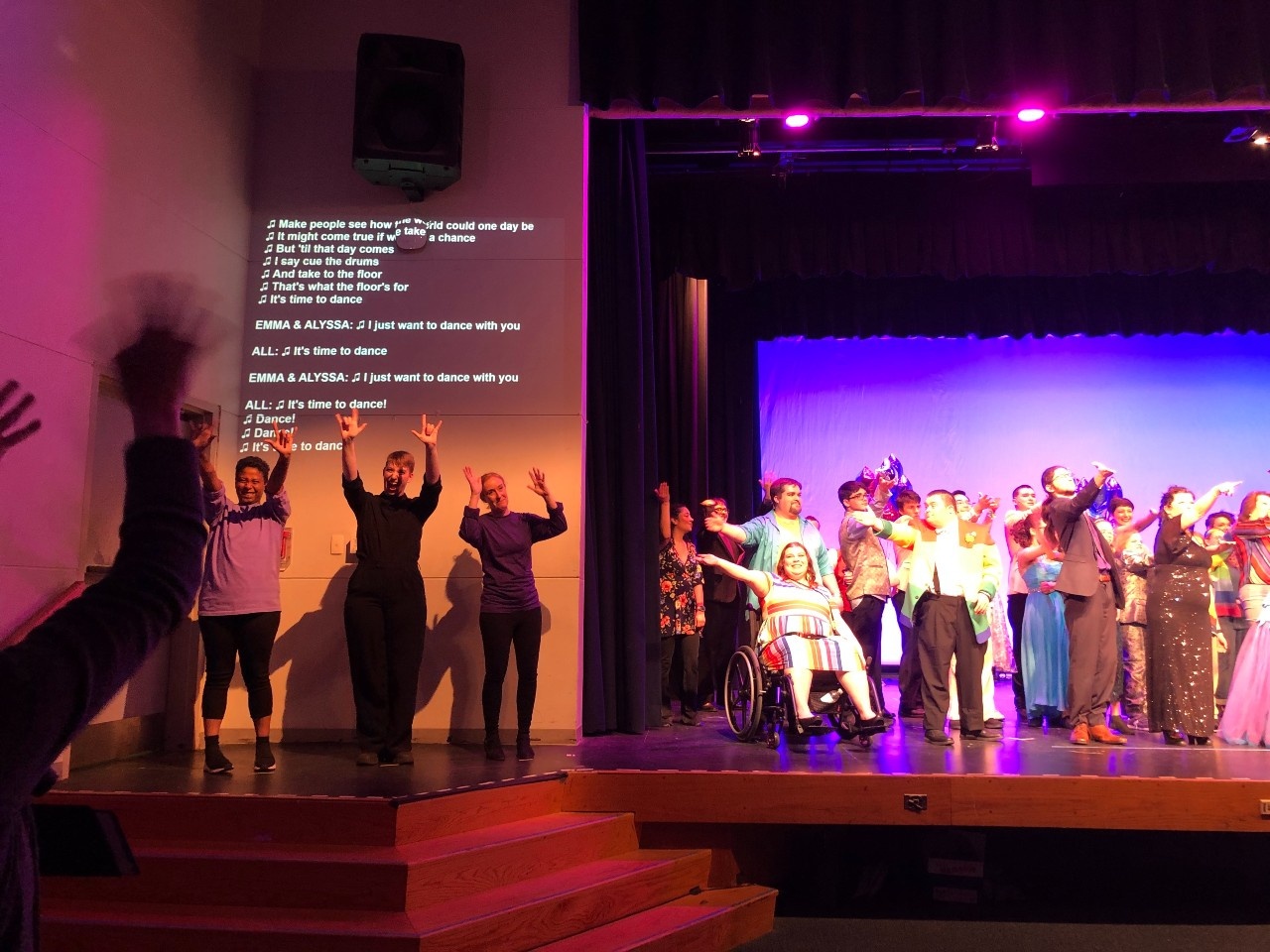The PROM cast bowing - they are in prom outfits which feature patterns, colors and sparkle. Some of the cast extend hands out to the audience, and on the left is the ASL team with hands waving and smiling, showing “I love you” hands to the crowd.