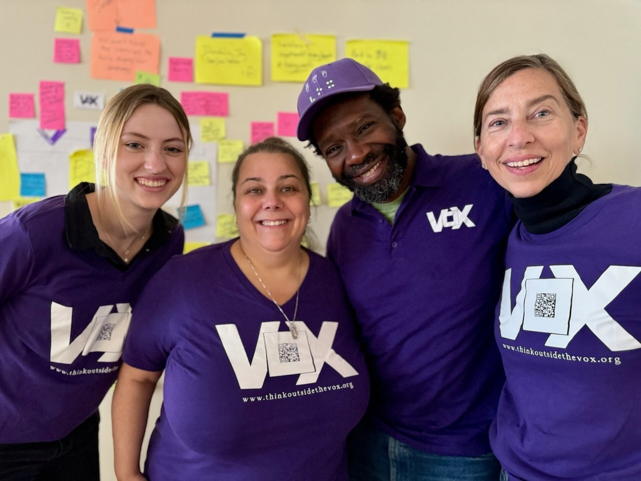 Vox team members standing and smiling with arms around each other wearing purple Vox shirts, jeans and dark shoes in front of a wall decorated with a paper map for a 2D organizational activity. From left to right: Ingrid O’Dell, a tall white woman with bl