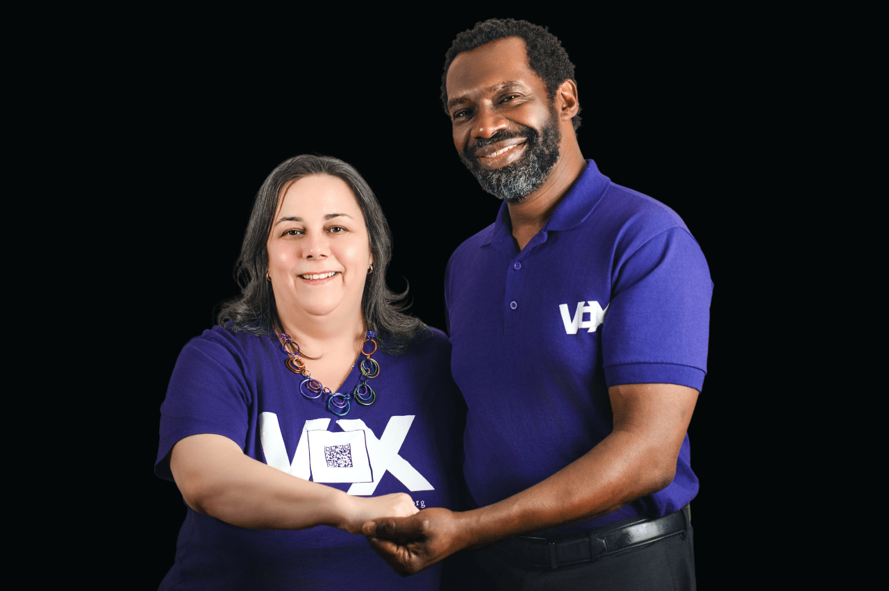 Sam and Chris in Vox purple with hands clasping to form the ASL sign for partner or collaborate.