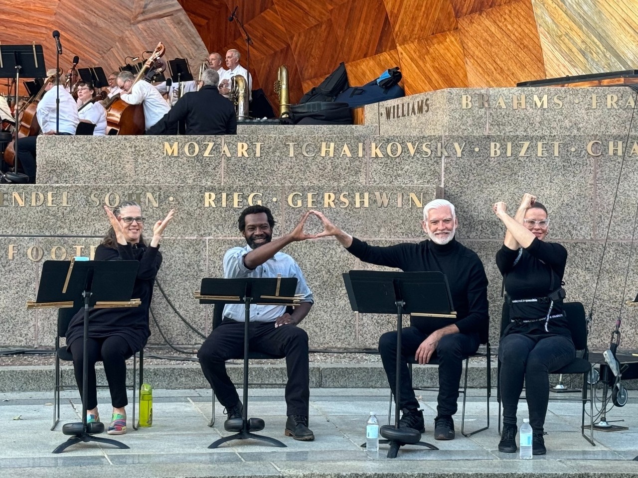 A photo of the ASL team at the Hatch Shell in front of the Landmarks Orchestra for the performance of Sheherazade & Borodin: Arabian Nights. There are stone steps with names of composers in gold: “Mozart, Tchaikovsky, Bizet, Brahms, Gershwin..” are among