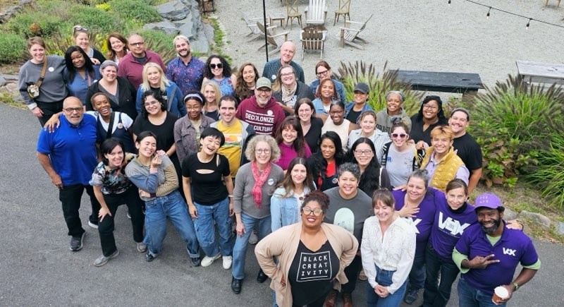 Large group photo taken from above of the NAS conference goers - a diverse group of BIPOC and white arts, access, and cultural workers standing in a driveway with an outdoor courtyard behind them. All are smiling, some are posing and some have arms around