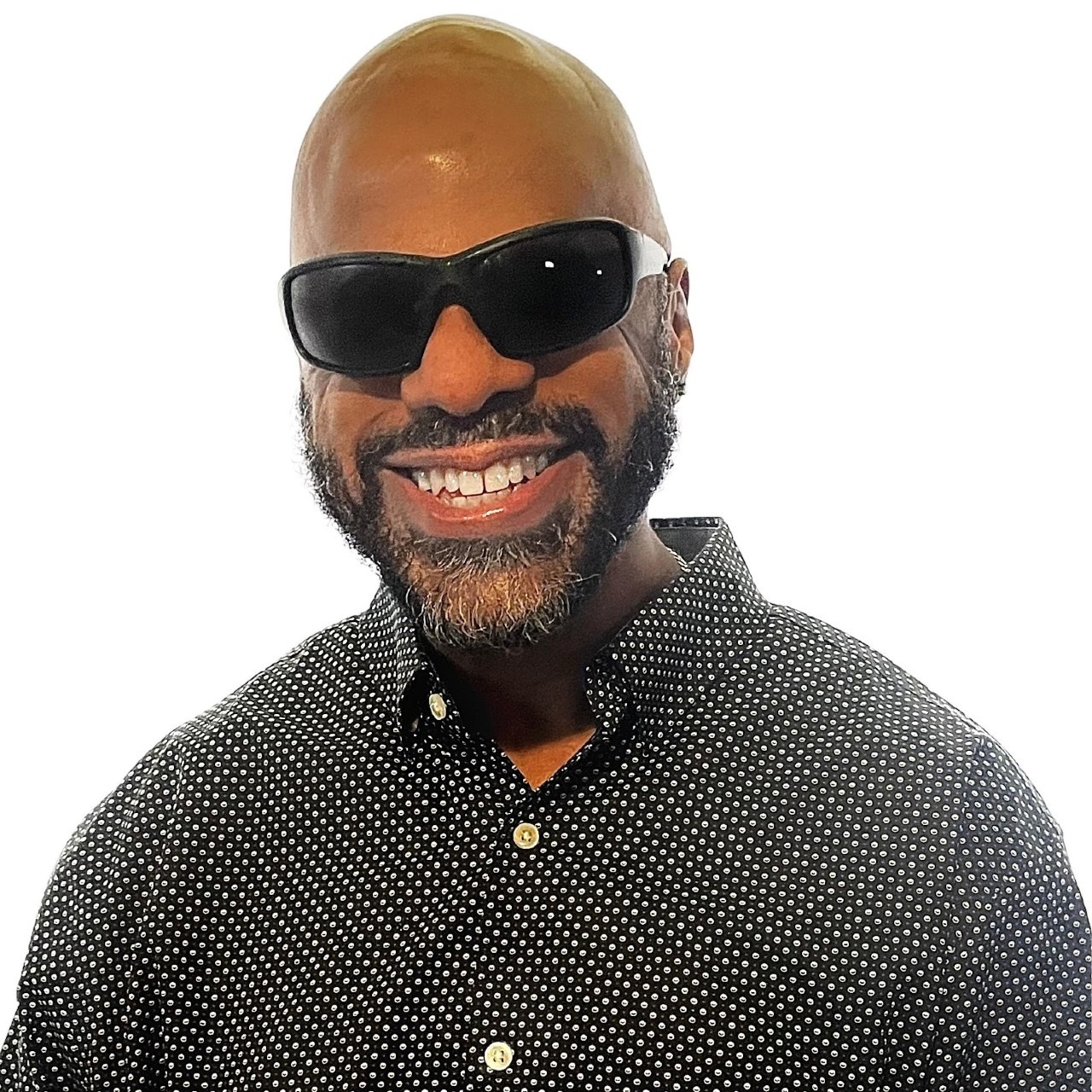 Thomas Reid, a brown skin Black man with a clean-shaven bald head and full neat beard, smiles into the camera. He wears dark shades and a black and white checkered button up shirt