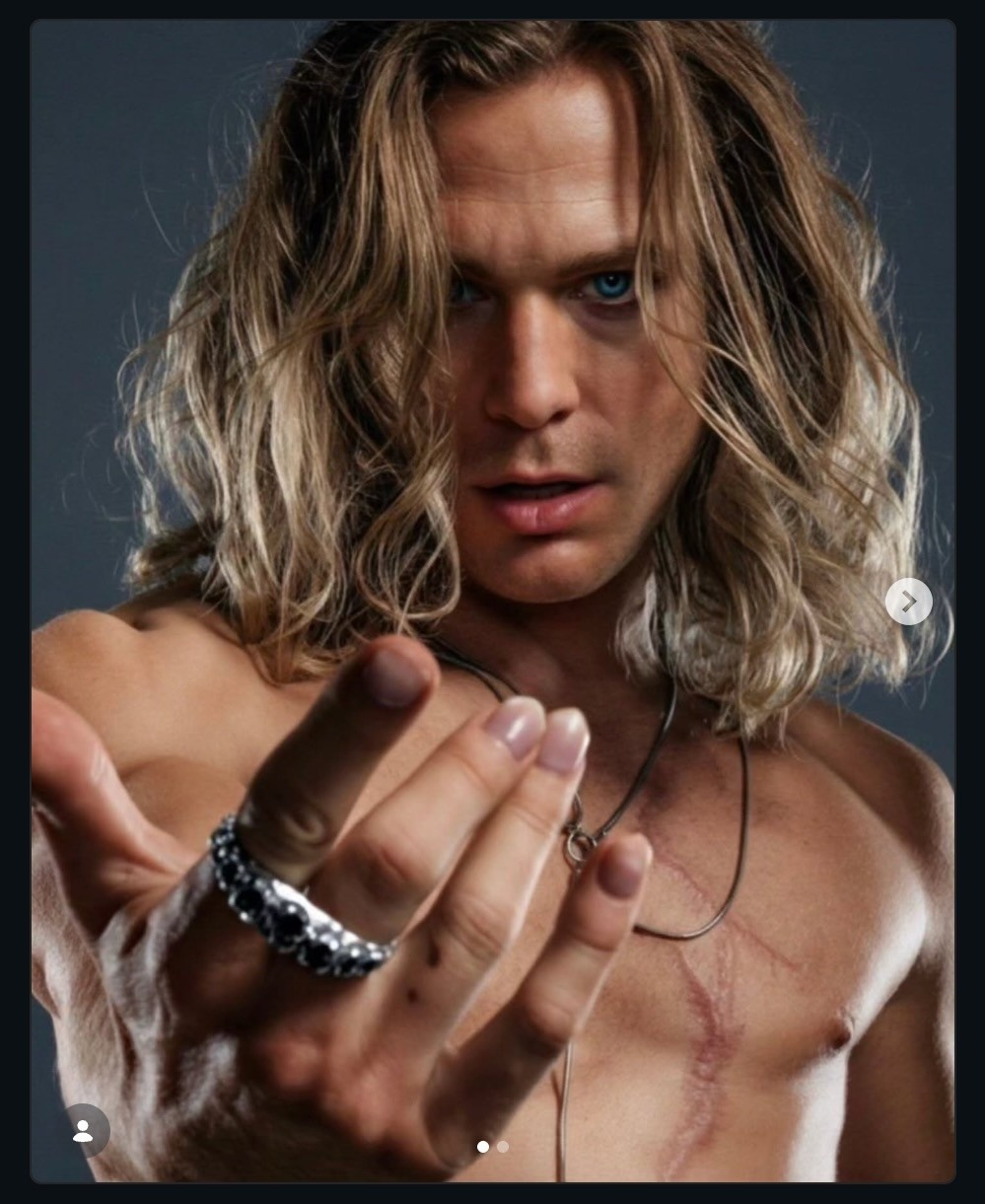 Image of Lestat with long, wavy hair and no shirt, holding his hand out towards the viewer with a seductive look on his face.