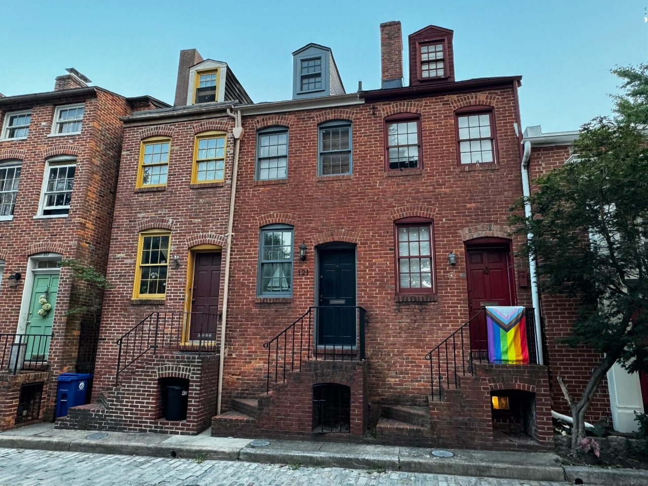 Photo of a street of small historic brick rowhouses in Otterbein. One of them is flying a rainbow progress flag.