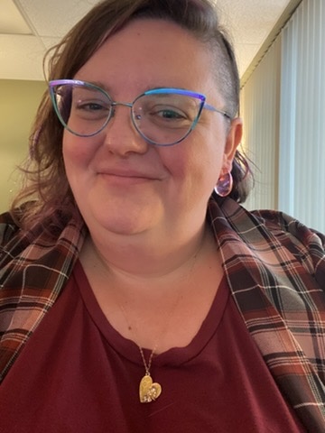 Jennifer Wolfe, owner of Kindness Corner Studio, wearing glasses and a red shirt, smiles warmly at the camera.