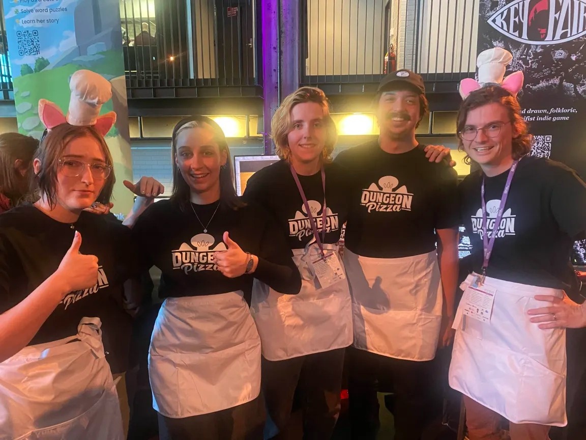 Image description: The Dungeon Pizza team posing for a group photo, with matching shirts, aprons - and Sam & Louis are wearing tiny chefs hats with cat ears.