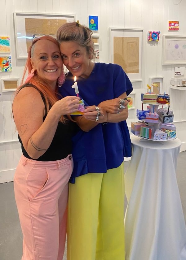 Fran and Morgane standing next to a cake stand with several decorated paper cake pieces. They are looking at the camera and smiling as they hold up one of the cake pieces with a lit candle on top.