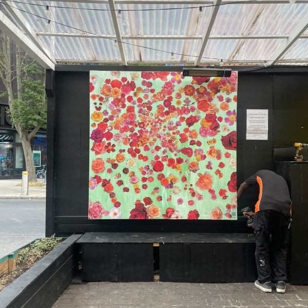 Our collaborative rose garden collage; glossy cut-out of blooming rose heads stuck on a painted green background. A man is screwing the stretched canvas onto a wall of the courtyard in Brixton Village.