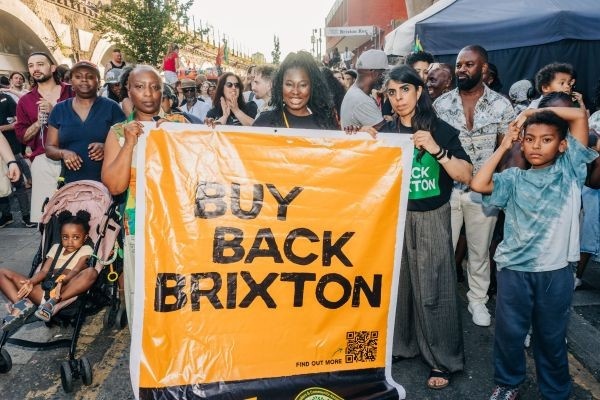 Kat, Akua, and Meera from the Brixton Traders Community Association holding their campaign poster. in front of a crowd of people gathered for the Buy Back Brixton Rally on Friday, the 19th of July 2026.