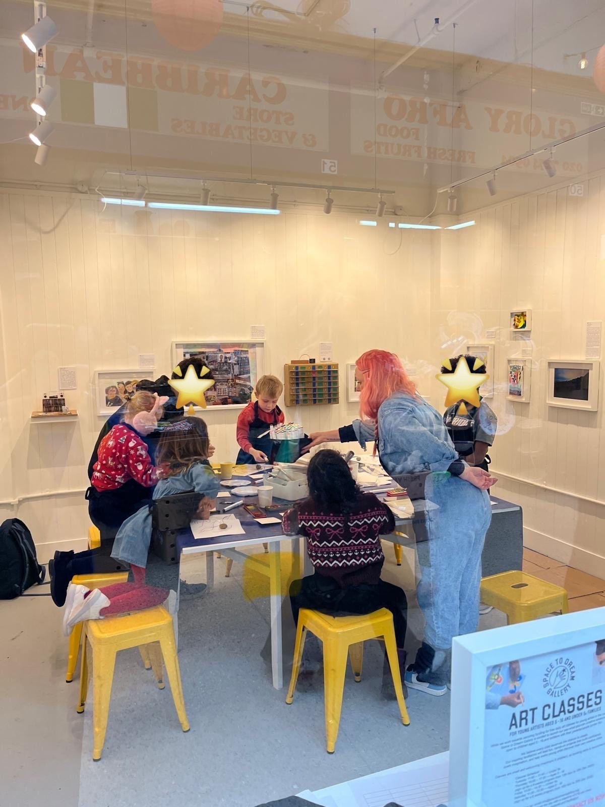 Photograph of one of our art classes in progress, taken from the outside of the gallery. It features a group of six children of primary school age involved in individual projects while Morgane, co-founder of the Space to Dream gallery, supports and guides