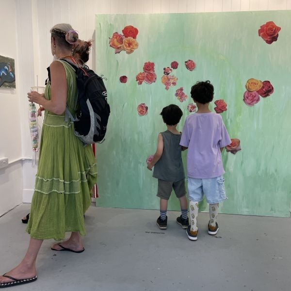 View inside the gallery with visitors on opening day of the CHANGE exhibition. While two women look at the art on display, two young boys stick paper flowers on the big collaborative garden collage.