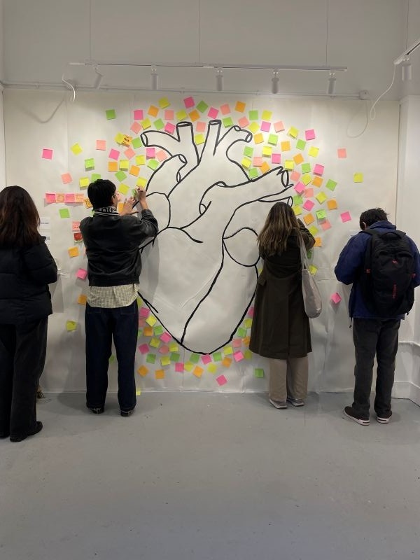 Photograph focusing on the back wall of the Space to Dream gallery, which features a giant drawing of an anatomical heart surrounded with multicoloured sticky notes. Some people are writing on notes to contribute to the display: What's in your Heart?