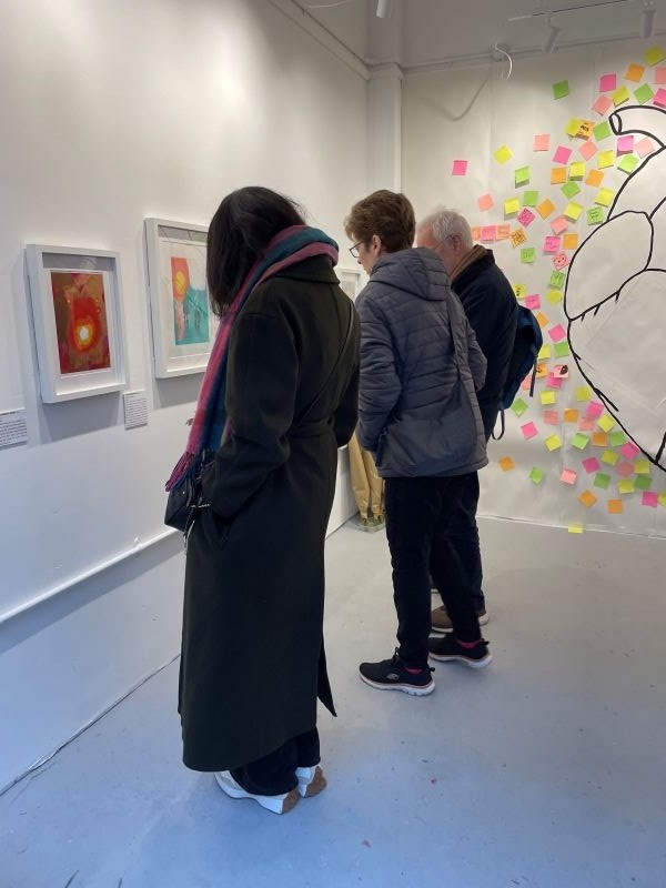 photograph taken at the Space to Dream gallery on the opening of the new exhibition; LOVE. The photograph features three people, two women and a man, looking at some of the work on display. We can also see part of the collaborative mural.