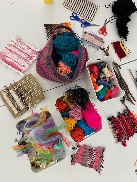 Photograph of an assortment of weaving materials on the table; wool, ribbons, scissors etc.