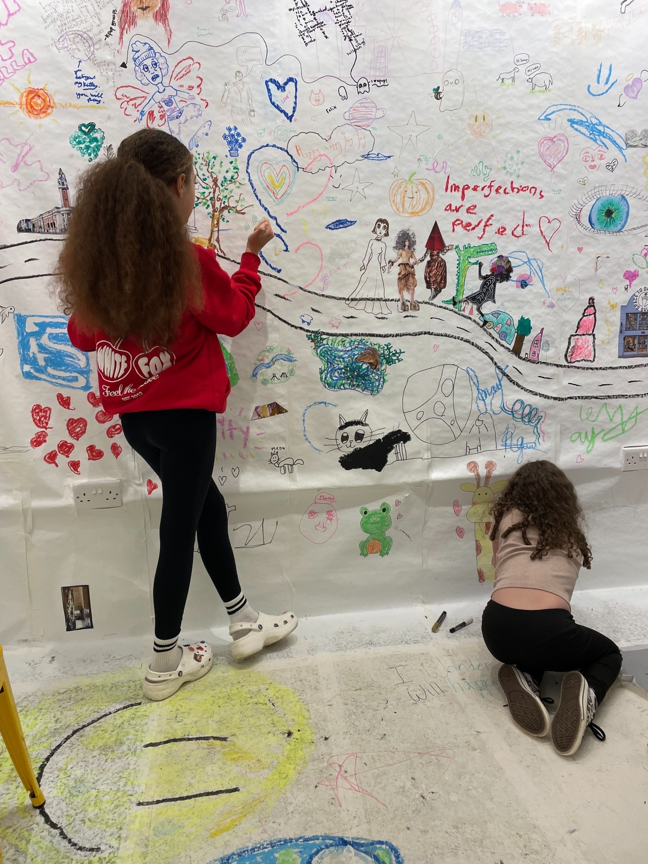 Two young girls of slightly different ages drawing on the wall. Part of the mural reads "Imperfections are Perfect", written in a child's handwriting.