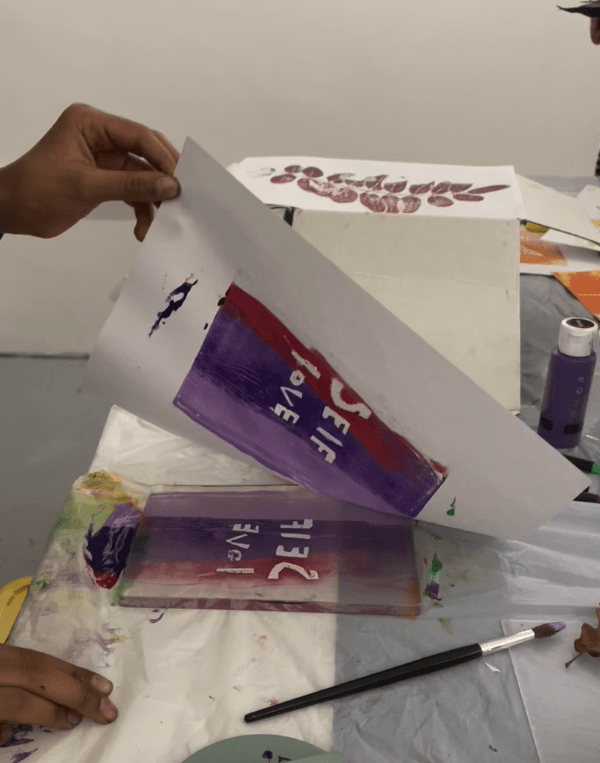 A close up of a young person's hand as they are lifting up a piece of paper from the block they have used to print it. Their design features a red and purple background with the words SELF LOVE appearing in white as they used cut-out letters.