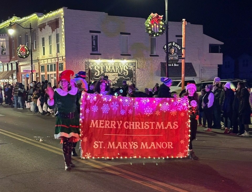 People dressed up for Christmas, holding a "Merry Christmas St. Marys Manor" banner while marching downtown St. Marys, KS