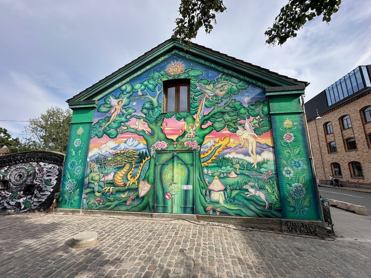 Building with elaborate fantastical mural on it, featuring nymphs, trees, elves, and other fairytale like beings, at the entrance to Christiania in Copenhagen