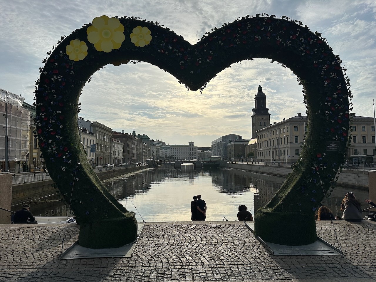 Heart-shaped gateway framing a couple hugging in front of a waterway with a steeple in the distance and obscured sunlight and dotted clouds with a cobblestone street in the foreground