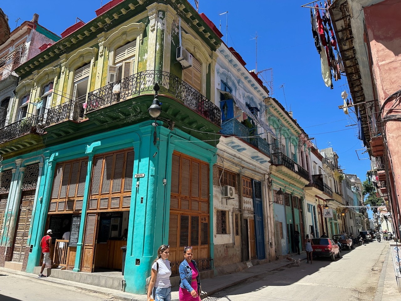 Colorful buildings with ornate facades and balconies in Havana with a few people walking on the dusty streets under a blue sky