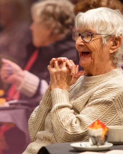 An elderly woman with short, grey hair and glasses, wearing a cream jumper sits in an audience looking delighted. She has a muffin and a cup next to her.