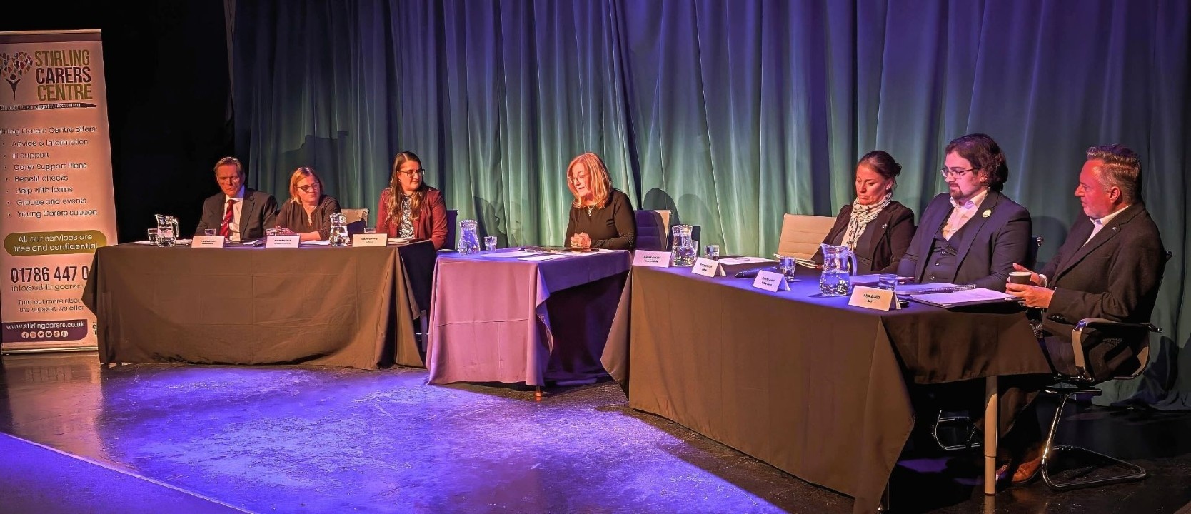 A group of six people sits at two tables on a stage in front of a blue curtain, prepared for a panel discussion.